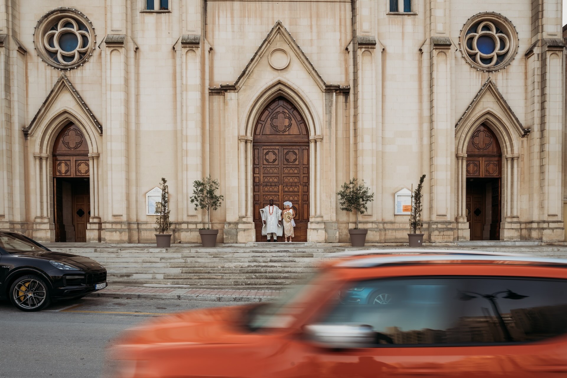 Bride and groom stand against a large, detailed wooden door as car whizzes by, destination wedding in Malta